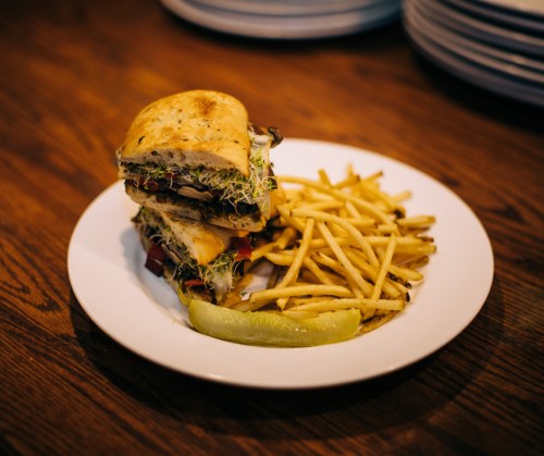 A stacked sandwich with sprouts, tomatoes, and pickles, served with a side of crispy fries on a white plate.