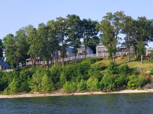 A serene lakeside view featuring a grassy hill with trees and houses, bordered by a white fence and a sandy shore.