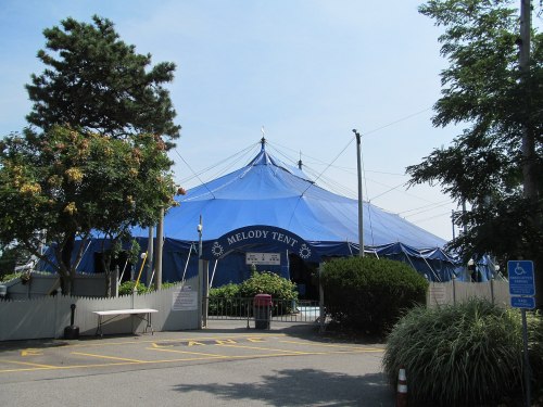A large blue circus tent labeled "Melody Tent" surrounded by trees and parking spaces on a sunny day.