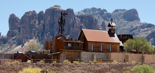 A rustic wooden church and buildings set against a backdrop of rugged mountains under a clear blue sky.