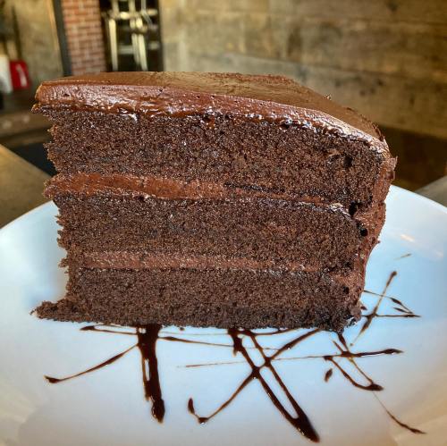 A slice of rich chocolate cake with layers of frosting, served on a white plate with chocolate drizzle.