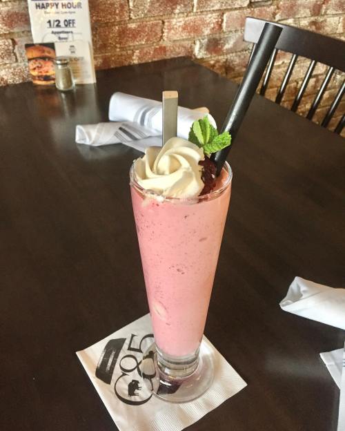 A tall glass of pink milkshake topped with whipped cream, a cherry, and a mint leaf, served on a napkin at a restaurant.
