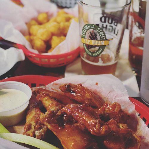 A basket of saucy chicken wings with celery and ranch, alongside a pint of beer and a plate of tater tots in the background.
