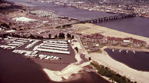 Aerial view of a river, marina, and bridge, with urban areas and green spaces in the background.