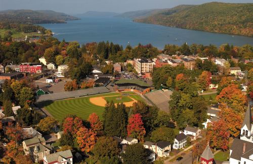 Aerial view of a town with a baseball field, colorful autumn trees, and a lake surrounded by hills in the background.
