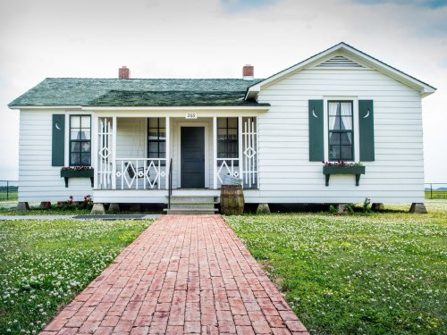 A white house with green shutters, a front porch, and a brick pathway surrounded by grass and flowers.
