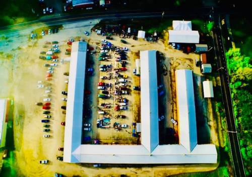 Aerial view of a large parking lot with rows of cars and several buildings, surrounded by trees and roads.