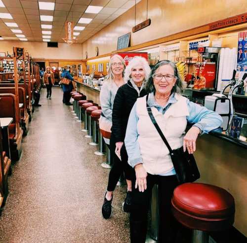 Three women pose at a diner counter with red stools, while others are seen in the background. Bright, nostalgic atmosphere.