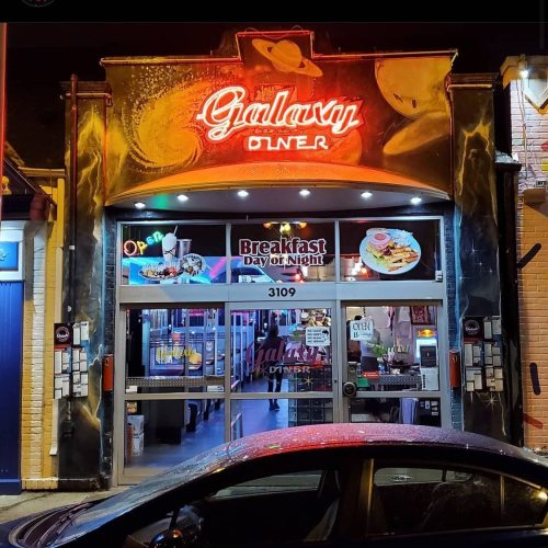 Brightly lit Galaxy Diner entrance at night, featuring a neon sign and large windows displaying breakfast options.