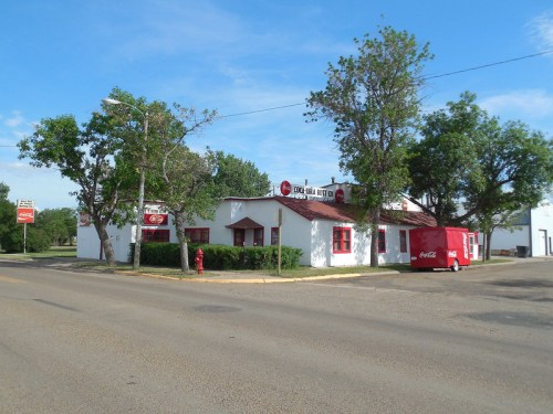 A white building with red accents and a Coca-Cola sign, surrounded by trees, on a quiet street.