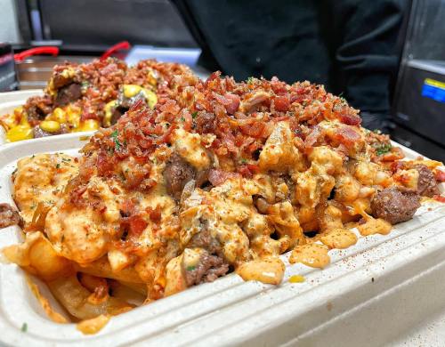 A close-up of loaded fries topped with cheese, bacon bits, and herbs in a takeout container.