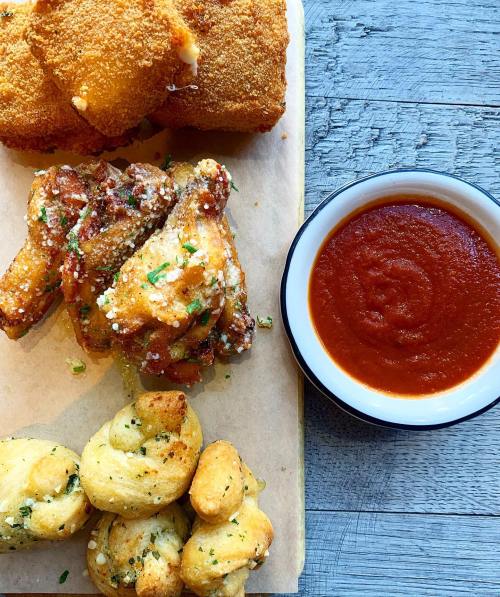 A wooden platter with crispy chicken wings, cheesy bites, and a bowl of marinara sauce.
