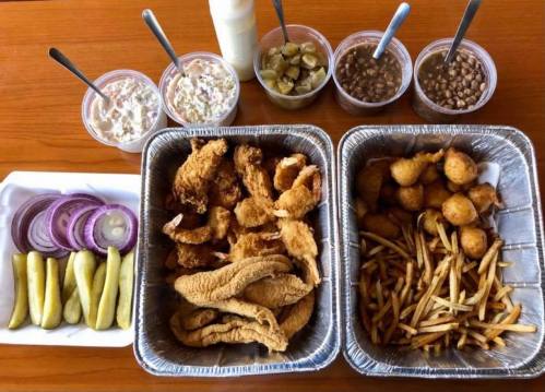 A spread of fried chicken, fish, pickles, onion slices, fries, and various desserts in cups on a wooden table.