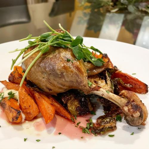 A plated dish featuring roasted duck leg atop a bed of caramelized carrots and Brussels sprouts, garnished with microgreens.