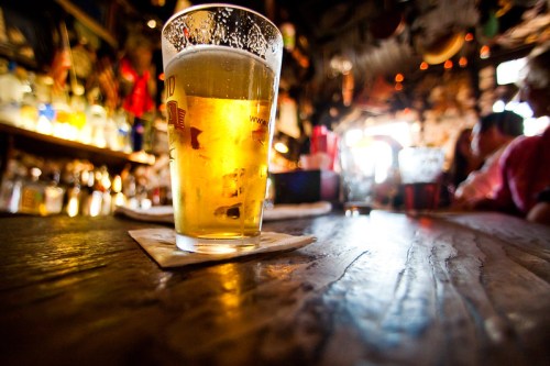 A close-up of a pint of beer on a wooden table, with a warm, lively bar atmosphere in the background.