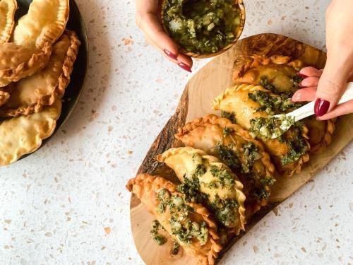 A wooden board with empanadas being topped with green sauce, hands holding a bowl of sauce, and a plate of empanadas nearby.