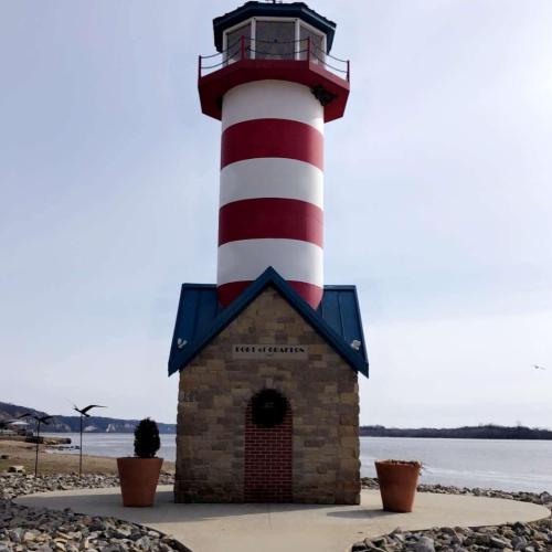 A red and white striped lighthouse with a blue roof, situated by a calm body of water and rocky shoreline.