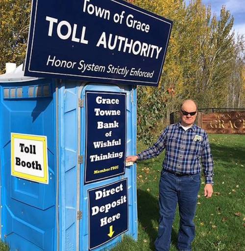 A man stands next to a humorous toll booth sign in Grace, featuring playful messages about a "bank of wishful thinking."