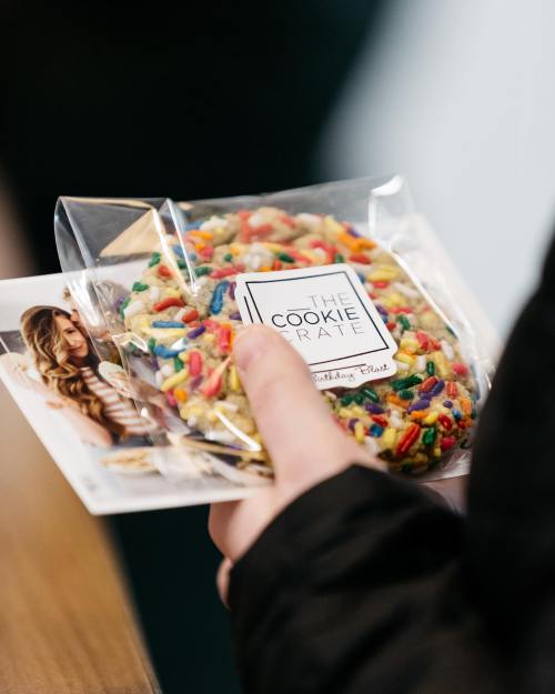 A hand holding a colorful cookie in a clear bag, next to a promotional card for "The Cookie Crate."