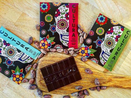 Three colorful chocolate bars with skull designs, surrounded by cocoa beans on a wooden board.