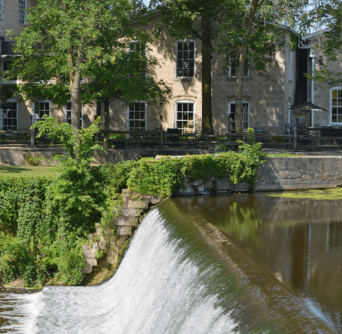 A serene waterfall cascades over a stone edge, surrounded by lush greenery and a historic building in the background.