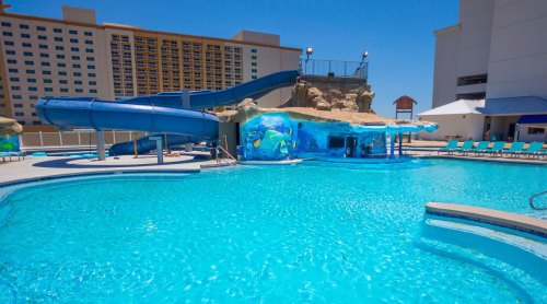 A bright blue pool with a water slide and a rock formation, surrounded by lounge chairs under a clear sky.