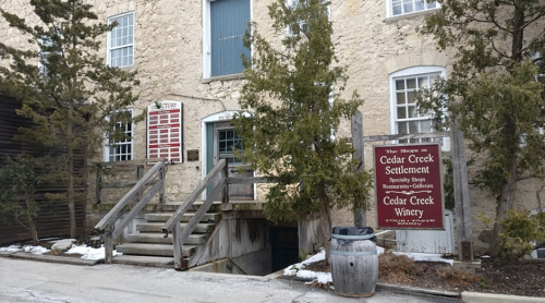 Exterior view of a stone building with a sign for Cedar Creek Winery and shops, surrounded by trees and snow.