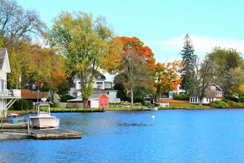 A serene lake scene with colorful autumn trees, houses along the shore, and a boat docked at a wooden pier.