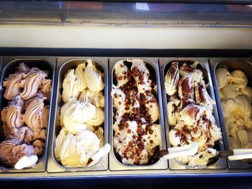 Four tubs of gelato in various flavors, including chocolate, vanilla, and a mix with toppings, displayed in a freezer.