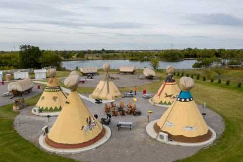 Aerial view of several colorful tipis near a lake, with people gathered around outdoor seating and picnic areas.