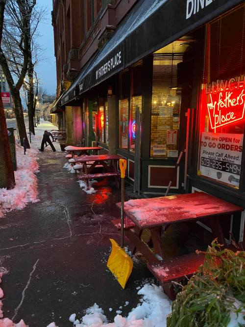 A snowy sidewalk in front of a diner with neon signs, featuring a shovel and a person clearing snow.