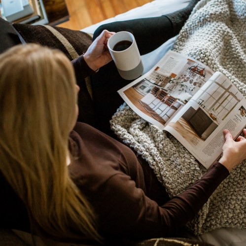A person sits on a bed, holding a coffee mug and reading a magazine, wrapped in a cozy blanket.