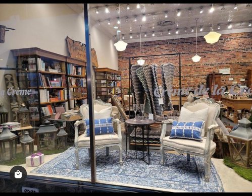 A cozy shop window display featuring two chairs, a table, bookshelves, and decorative items in a warm, inviting setting.