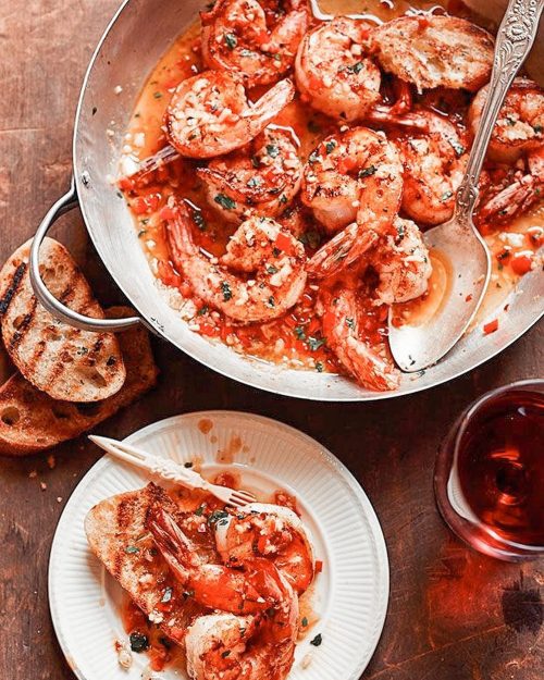 A skillet of shrimp in sauce, served with toasted bread on a plate and a glass of red drink nearby.