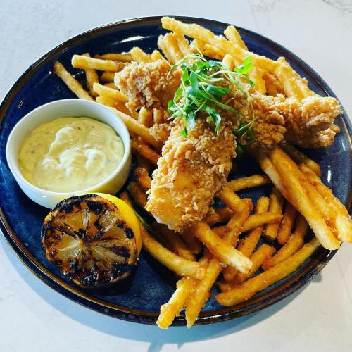 A plate of crispy fried chicken strips with French fries, a side of dipping sauce, and a grilled lemon.