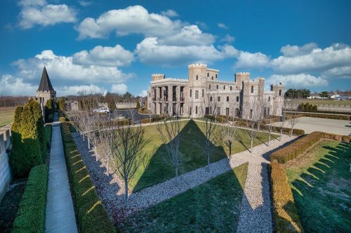 A large stone castle surrounded by manicured lawns and trees under a blue sky with fluffy clouds.