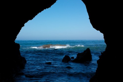 View of the ocean through a rocky cave, with waves gently crashing against the shore under a clear blue sky.