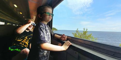 Two children look out a train window at a scenic view of the water and shoreline on a sunny day.