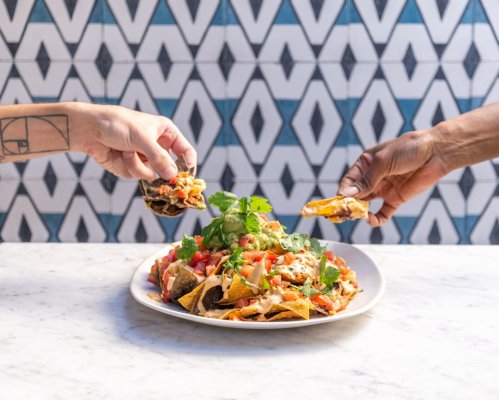 Two hands reaching for a plate of nachos topped with guacamole, tomatoes, and cilantro, set against a patterned background.