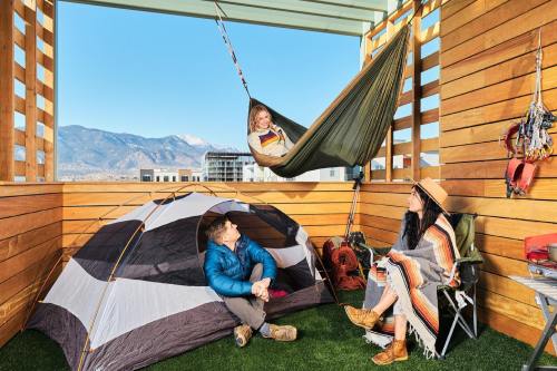 A cozy outdoor scene with a tent, hammock, and two people relaxing on a balcony with mountains in the background.