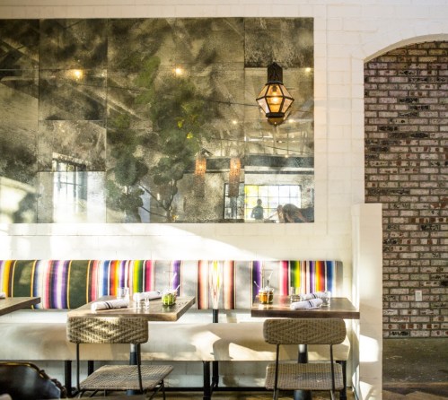 Bright restaurant interior featuring a striped banquette, wooden tables, and a large mirror with greenery.