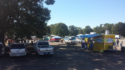 A bustling outdoor market scene with food trucks, tents, and parked cars under a clear blue sky.