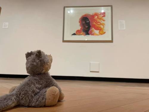 A stuffed animal gazes at a colorful portrait of a woman with fiery hair in an art gallery.