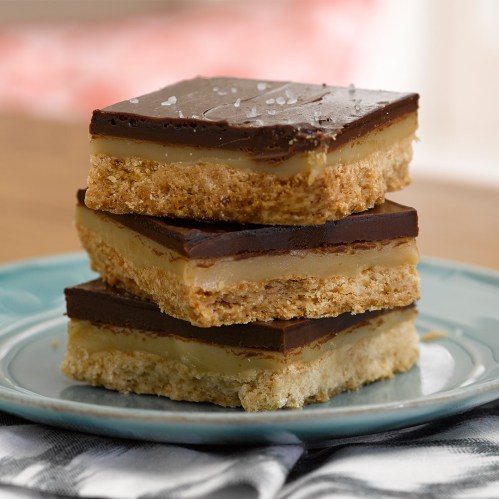 Three layered dessert bars with a chocolate topping, creamy filling, and a crumbly base, served on a blue plate.