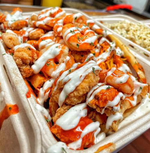 A close-up of a takeout container filled with shrimp, fries, and drizzled with sauce and ranch dressing.