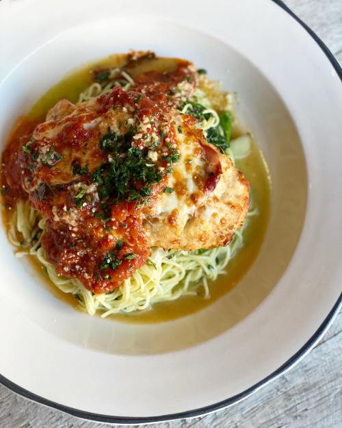 A plate of pasta topped with marinara sauce, herbs, and a golden-brown chicken breast, served with greens.