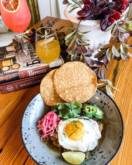 A plate of food featuring a fried egg, tortillas, pickled onions, jalapeños, and lime, with drinks and books in the background.