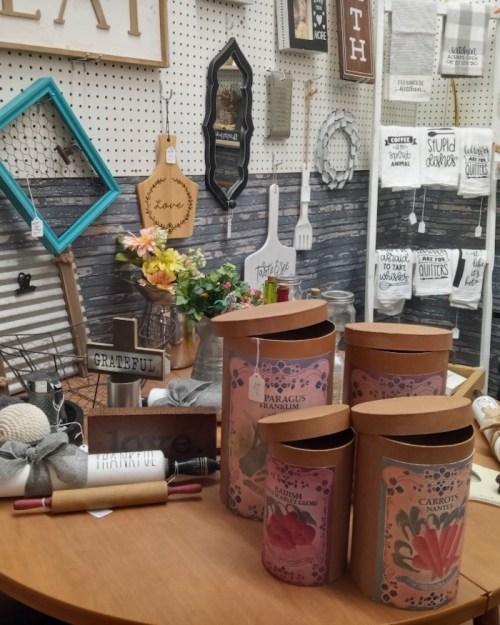 A display of decorative items, including jars, signs, and flowers, arranged on a table against a textured wall.