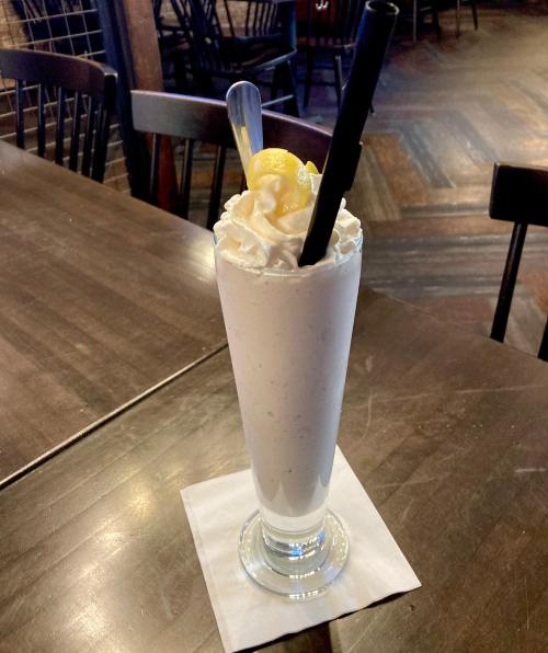 A tall glass of creamy milkshake topped with whipped cream and a lemon slice, sitting on a wooden table.
