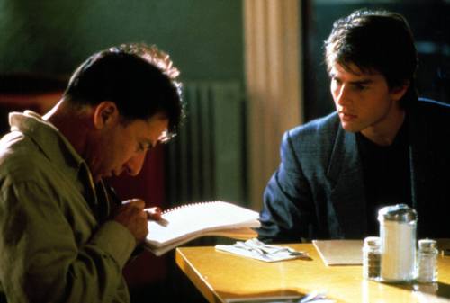 Two men sit at a table, one writing in a notebook while the other observes intently, in a dimly lit room.